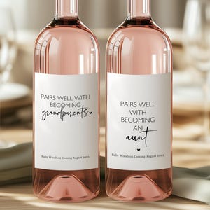 May include: Two bottles of rosé wine with white labels. One label reads "Pairs well with becoming grandparents" and the other reads "Pairs well with becoming an aunt". The bottles have a pink hue and are displayed on a wooden table.