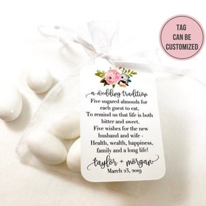 Puede incluir: Una bolsa de organza blanca llena de cinco almendras azucaradas blancas. Una etiqueta blanca con un dise&ntilde;o floral y el texto "a wedding tradition" est&aacute; unida a la bolsa. La etiqueta tambi&eacute;n incluye el texto "Five sugared almonds for each guest to eat, To remind us that life is both bitter and sweet. Five wishes for the new husband and wife - Health, wealth, happiness, family and a long life! taylor + morgan March 23, 2019". Un c&iacute;rculo rosa con el texto "TAG CAN BE CUSTOMIZED" est&aacute; en la esquina superior derecha de la imagen.