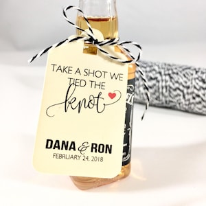 May include: A cream-colored tag with black text that reads "Take a shot we tied the knot" with a red heart. The tag also has the names "Dana & Ron" and the date "February 24, 2018".