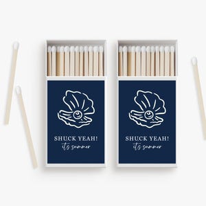 May include: Two matchboxes with navy blue covers featuring a white seashell design and the text "SHUCK YEAH! it's summer." The boxes are open, revealing wooden matches with white tips. Two loose matches are also visible.