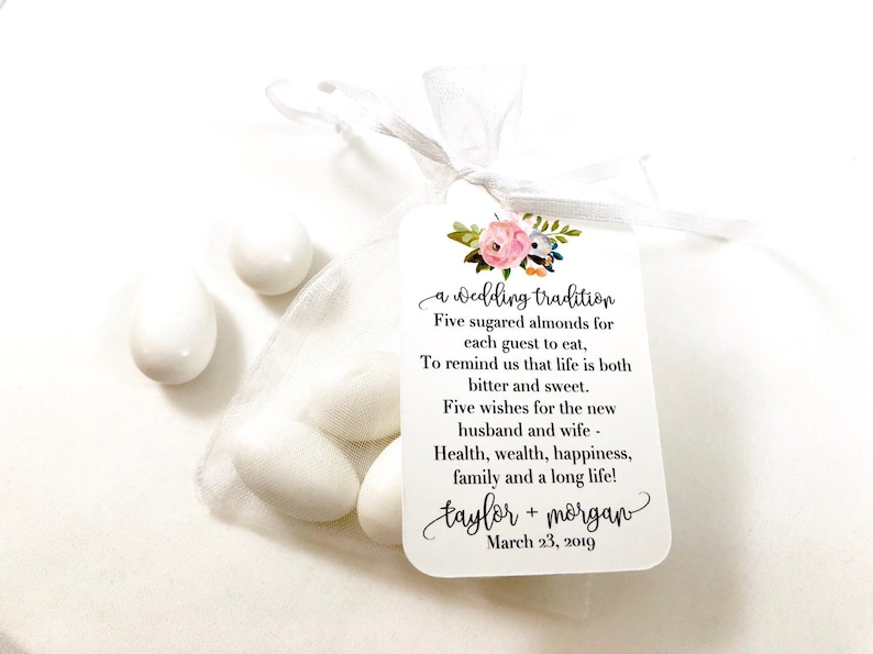 Puede incluir: Una bolsa de organza blanca llena de cinco almendras azucaradas blancas. Una etiqueta blanca con detalles florales rosas y texto negro dice "a wedding tradition Five sugared almonds for each guest to eat, To remind us that life is both bitter and sweet. Five wishes for the new husband and wife - Health, wealth, happiness, family and a long life! taylor + morgan March 23, 2019"