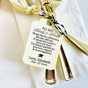 May include: A white tag with black text that reads "To My Family & Friends" and includes a graduation cap icon. The text says "Thank you for celebrating this day with me. It means the world to have you here! I'm forever grateful for your love, support & encouragement. Love, Elizabeth Class of 20xx". The tag is tied to two gold spoons.