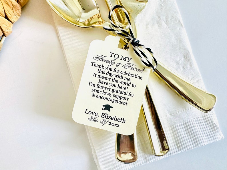 May include: A white tag with black text that reads "To My Family & Friends" and includes a graduation cap icon. The text says "Thank you for celebrating this day with me. It means the world to have you here! I'm forever grateful for your love, support & encouragement. Love, Elizabeth Class of 20xx". The tag is tied to two gold spoons.
