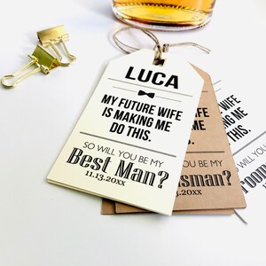 May include: Three white and brown gift tags with black text. The top tag says "LUCA" with a bow tie and "MY FUTURE WIFE IS MAKING ME DO THIS. SO WILL YOU BE MY BEST MAN? 11.13.20XX". The middle tag says "SO WILL YOU BE MY BEST MAN? 11.13.20XX". The bottom tag says "SO WILL YOU BE MY GROOMSMAN? 11.13.20XX".