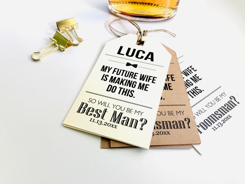 May include: Three white and brown gift tags with black text. The top tag says "LUCA" with a bow tie and "MY FUTURE WIFE IS MAKING ME DO THIS. SO WILL YOU BE MY BEST MAN? 11.13.20XX". The middle tag says "SO WILL YOU BE MY BEST MAN? 11.13.20XX". The bottom tag says "SO WILL YOU BE MY GROOMSMAN? 11.13.20XX".