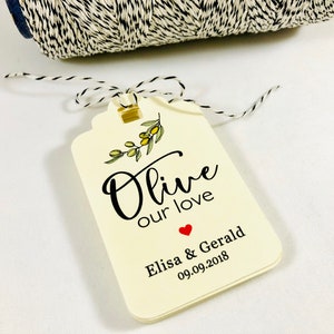 May include: A white gift tag with a black and green olive branch illustration and the text "Olive our love" with a red heart and the names "Elisa & Gerald" and the date "09.09.2018".
