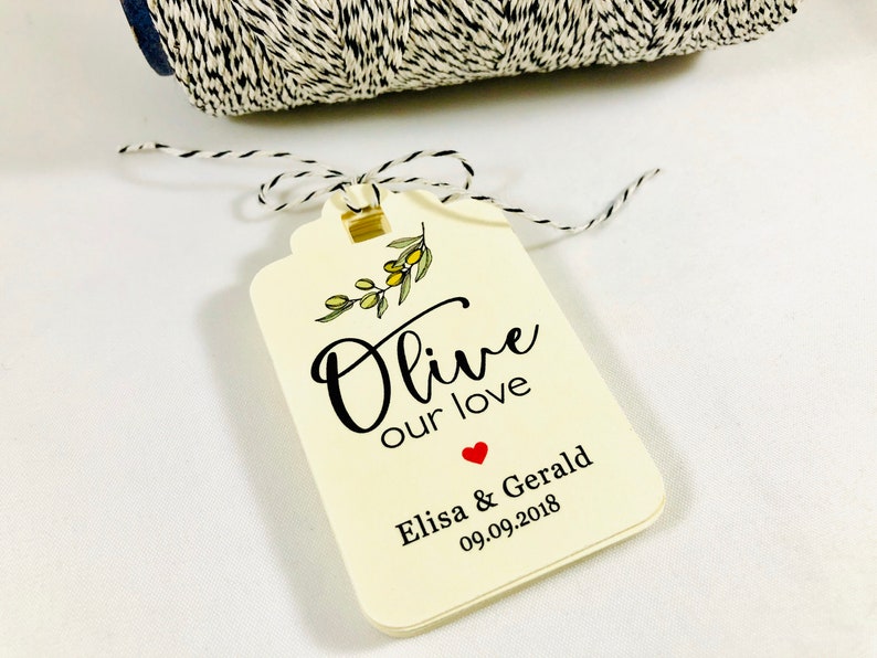 May include: A white gift tag with a black and green olive branch illustration and the text "Olive our love" with a red heart and the names "Elisa & Gerald" and the date "09.09.2018".