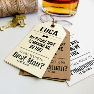 May include: Three white, brown, and cream paper gift tags with black text. The top tag reads "LUCA MY FUTURE WIFE IS MAKING ME DO THIS. SO WILL YOU BE MY BEST MAN? 11.13.20XX". The middle tag reads "SO WILL YOU BE MY BEST MAN? 11.13.20XX". The bottom tag reads "SO WILL YOU BE MY GROOMSMAN? 11.13.20XX".