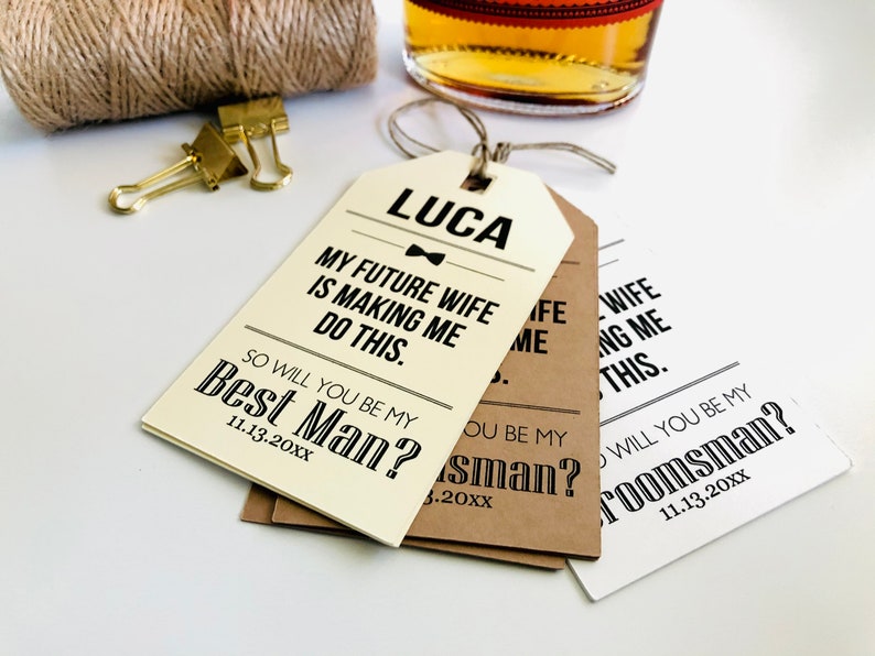 May include: Three white, brown, and cream paper gift tags with black text. The top tag reads "LUCA MY FUTURE WIFE IS MAKING ME DO THIS. SO WILL YOU BE MY BEST MAN? 11.13.20XX". The middle tag reads "SO WILL YOU BE MY BEST MAN? 11.13.20XX". The bottom tag reads "SO WILL YOU BE MY GROOMSMAN? 11.13.20XX".