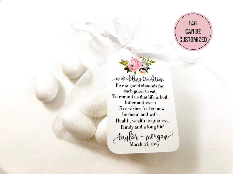 Puede incluir: Una bolsa de organza blanca llena de cinco almendras azucaradas blancas. Una etiqueta blanca con un dise&ntilde;o floral y el texto "a wedding tradition" est&aacute; unida a la bolsa. La etiqueta tambi&eacute;n incluye el texto "Five sugared almonds for each guest to eat, To remind us that life is both bitter and sweet. Five wishes for the new husband and wife - Health, wealth, happiness, family and a long life! taylor + morgan March 23, 2019". Un c&iacute;rculo rosa con el texto "TAG CAN BE CUSTOMIZED" est&aacute; en la esquina superior derecha de la imagen.