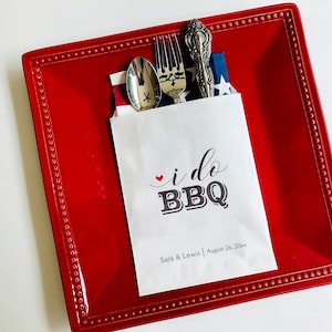 May include: White paper bag with the text "i do BBQ" in black and a red heart. The bag is on a red plate with a white napkin and silverware.