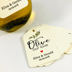 May include: A stack of ivory-colored gift tags with a small olive branch illustration and the text "Olive our love" in black script. The bottom tag has the text "Elisa & Gerald 09.09.2018" in black text with a small red heart.