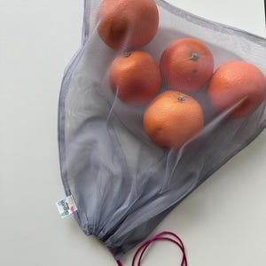 Op de afbeelding: Een grijze mesh-tas voor producten met een roze trekkoord, gevuld met vijf oranje sinaasappelen. De tas heeft een klein label met de tekst "Love Your Planet".