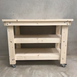 May include: A natural wood workbench with three shelves and wheels. The workbench is made of unfinished pine wood and has a simple design.
