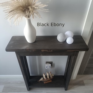 Op de afbeelding: Een donker gebeitste houten consoletafel met een onderste plank. Een witte vaas met gedroogd siergras staat op de tafel, samen met drie decoratieve bollen. De tekst "Black Ebony" is zichtbaar op de afbeelding. Een klein houten scheepsmodel staat op de onderste plank.