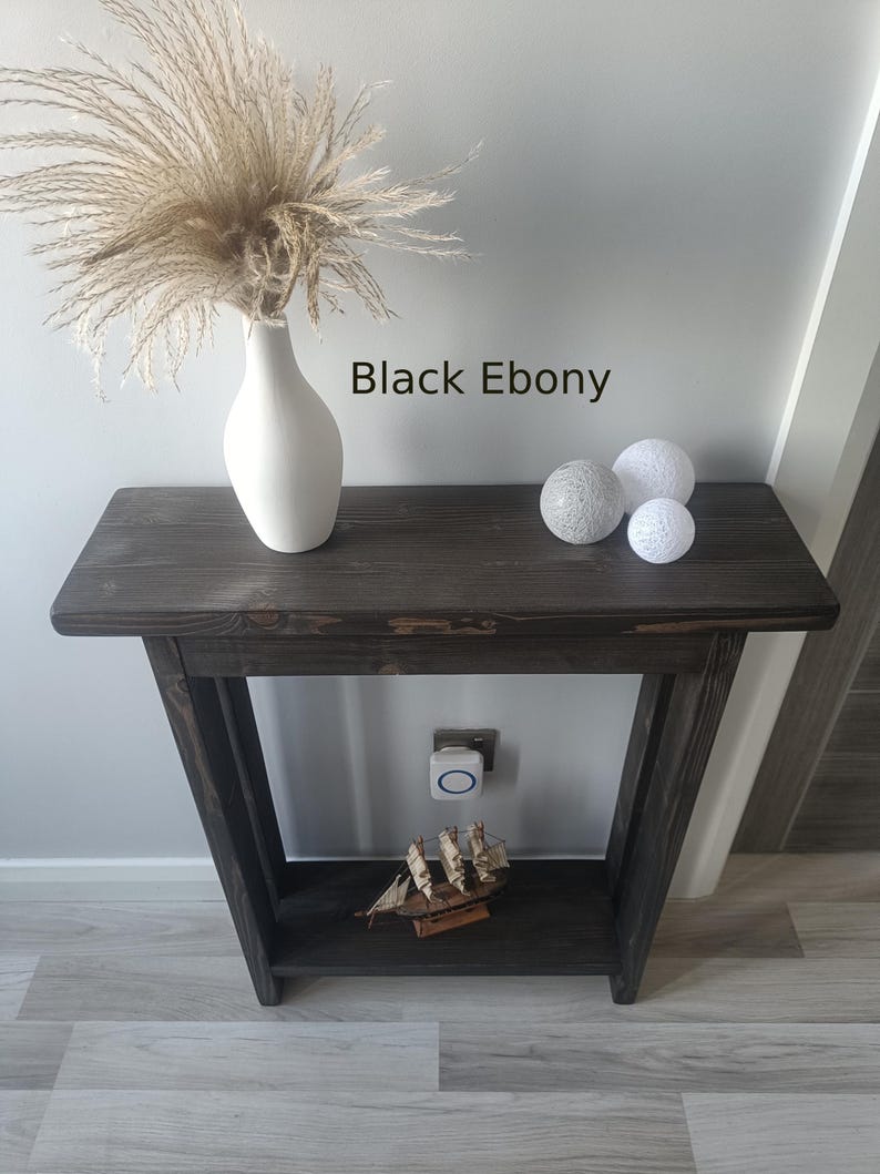 Op de afbeelding: Een donker gebeitste houten consoletafel met een onderste plank. Een witte vaas met gedroogd siergras staat op de tafel, samen met drie decoratieve bollen. De tekst "Black Ebony" is zichtbaar op de afbeelding. Een klein houten scheepsmodel staat op de onderste plank.