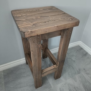 K&ouml;nnte beinhalten: Ein rustikaler, quadratischer Holzschemel mit nat&uuml;rlicher Oberfl&auml;che. Der Hocker hat eine quadratische Oberseite und vier stabile Beine, die die Holzmaserung zeigen. Der Hocker ist etwa 61 cm hoch.