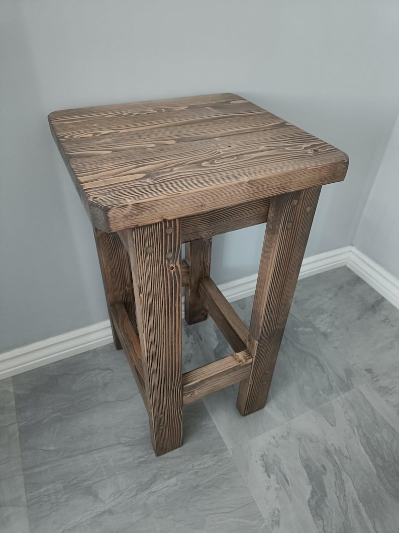 K&ouml;nnte beinhalten: Ein rustikaler, quadratischer Holzschemel mit nat&uuml;rlicher Oberfl&auml;che. Der Hocker hat eine quadratische Oberseite und vier stabile Beine, die die Holzmaserung zeigen. Der Hocker ist etwa 61 cm hoch.