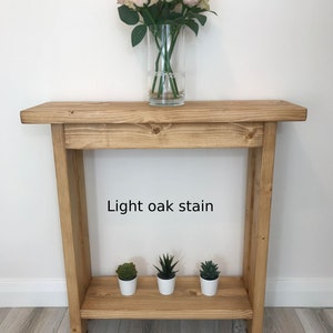Op de afbeelding: Een licht eikenhouten console met een onderste plank. Bovenop staat een heldere glazen vaas met roze rozen. Drie kleine vetplanten in potten staan op de onderste plank. De tekst "Light oak stain" is zichtbaar.