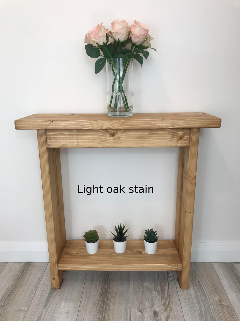 Op de afbeelding: Een licht eikenhouten console met een onderste plank. Bovenop staat een heldere glazen vaas met roze rozen. Drie kleine vetplanten in potten staan op de onderste plank. De tekst "Light oak stain" is zichtbaar.