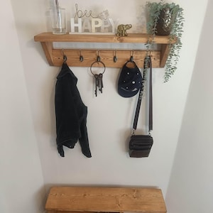 May include: A wall-mounted wooden coat rack with five black metal hooks, featuring a top shelf displaying a glass vase, a sign reading "bee HAPPY", a small gold elephant figurine, and a potted plant. Below sits a matching wooden shoe bench.