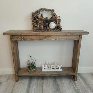 Op de afbeelding: Een rustieke houten console met een onderste plank. Bovenop staat een decoratieve klok met figuren. De onderste plank bevat een kleine hangende plantenbak met groen en een wit bord met de tekst "bee HAPPY".