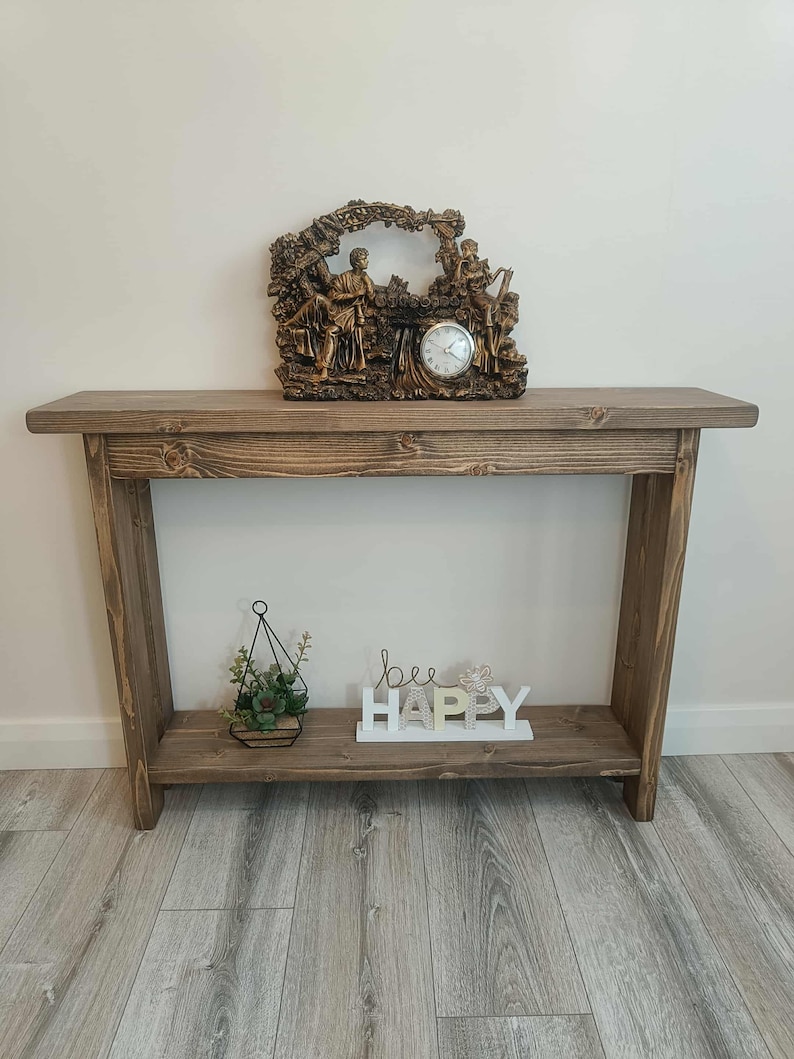 Op de afbeelding: Een rustieke houten console met een onderste plank. Bovenop staat een decoratieve klok met figuren. De onderste plank bevat een kleine hangende plantenbak met groen en een wit bord met de tekst "bee HAPPY".