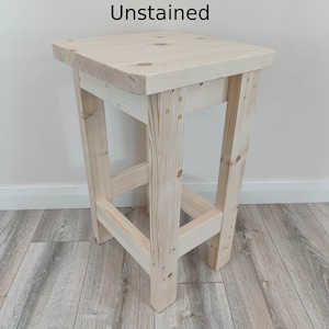 K&ouml;nnte beinhalten: Ein unbehandelter Holzstuhl mit quadratischer Sitzfl&auml;che und vier abgewinkelten Beinen. Der Text "Unstained" ist auf der Oberseite aufgedruckt. Der Hocker besteht aus Naturholz und weist sichtbare Maserungen auf. Die H&ouml;he betr&auml;gt ca. 76 cm.