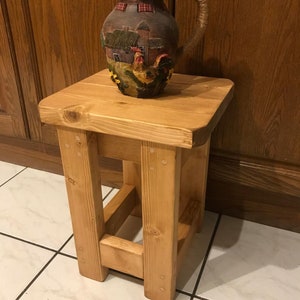 K&ouml;nnte beinhalten: Ein kleiner, hellfarbiger Holzschemel mit quadratischer Oberseite und vier stabilen Beinen. Auf der Oberseite steht ein dekorativer Keramikkrug mit einer Bauernhofszene und einem Hahn-Design. Der Krug hat einen griff aus Seil.