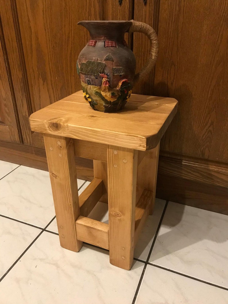 K&ouml;nnte beinhalten: Ein kleiner, hellfarbiger Holzschemel mit quadratischer Oberseite und vier stabilen Beinen. Auf der Oberseite steht ein dekorativer Keramikkrug mit einer Bauernhofszene und einem Hahn-Design. Der Krug hat einen griff aus Seil.
