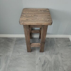 K&ouml;nnte beinhalten: Ein rustikaler Holzschemel mit quadratischer Sitzfl&auml;che und vier stabilen Beinen. Das Holz hat eine nat&uuml;rliche Maserung und einen warmen braunen Farbton. Der Hocker ist mit einer einfachen, funktionalen &Auml;sthetik gestaltet, passend f&uuml;r verschiedene Innenr&auml;ume. H&ouml;he: ca. 45 cm.