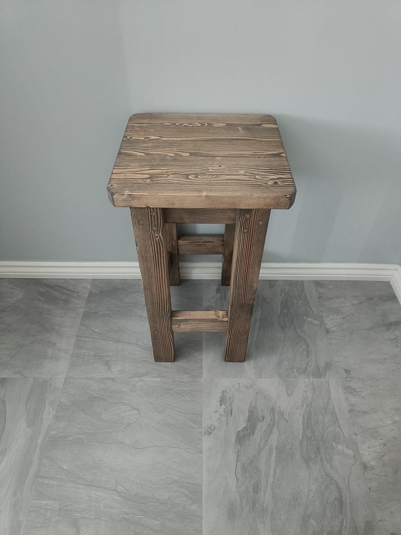 K&ouml;nnte beinhalten: Ein rustikaler Holzschemel mit quadratischer Sitzfl&auml;che und vier stabilen Beinen. Das Holz hat eine nat&uuml;rliche Maserung und einen warmen braunen Farbton. Der Hocker ist mit einer einfachen, funktionalen &Auml;sthetik gestaltet, passend f&uuml;r verschiedene Innenr&auml;ume. H&ouml;he: ca. 45 cm.