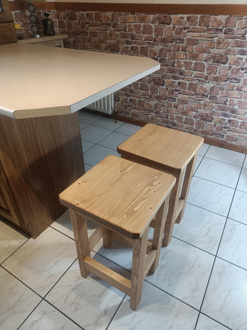 K&ouml;nnte beinhalten: Zwei Holzst&uuml;hle mit nat&uuml;rlicher Oberfl&auml;che. Die Hocker haben eine quadratische Sitzfl&auml;che und vier stabile Beine. Die Holzmaserung ist sichtbar, was ihnen ein rustikales Aussehen verleiht. Sie stehen vor einer K&uuml;chenzeile.
