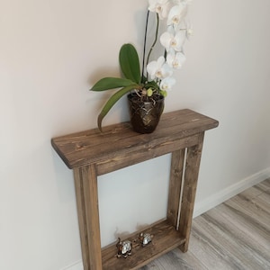Op de afbeelding: Een rustieke houten console met een onderste plank. Bovenop staat een orchidee in pot met witte bloemen en groene bladeren. Twee kleine decoratieve beeldjes staan op de onderste plank. De tafel is gemaakt van gebeitst hout.
