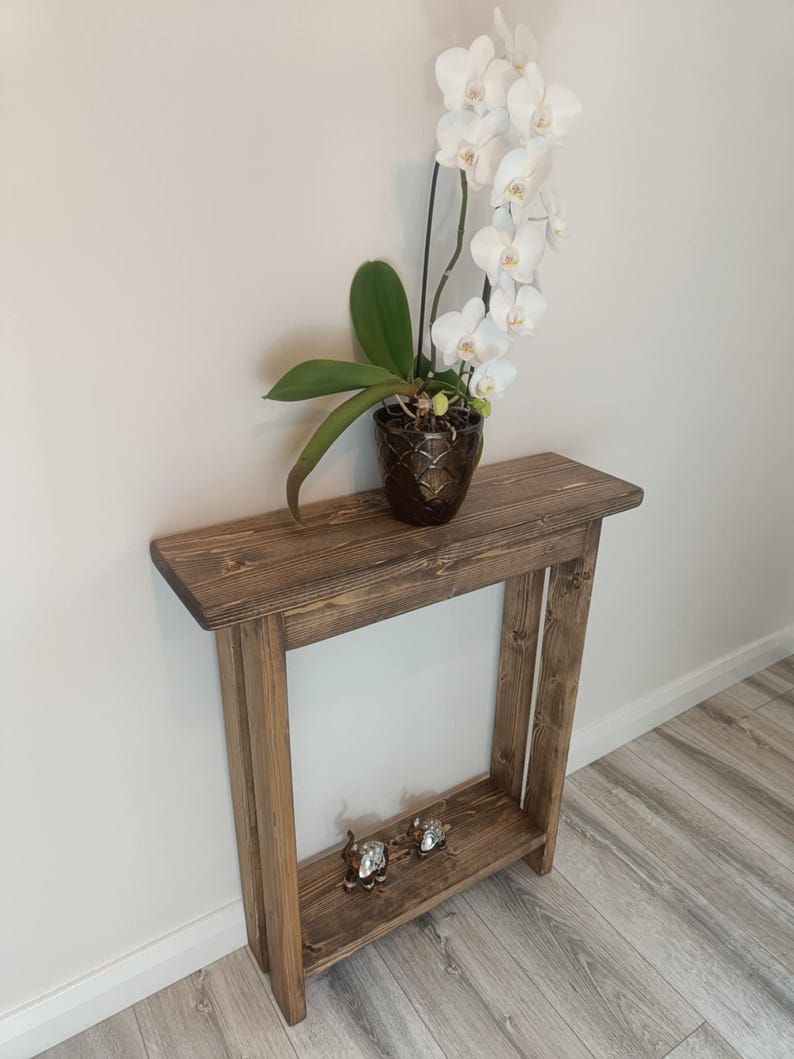 Op de afbeelding: Een rustieke houten console met een onderste plank. Bovenop staat een orchidee in pot met witte bloemen en groene bladeren. Twee kleine decoratieve beeldjes staan op de onderste plank. De tafel is gemaakt van gebeitst hout.