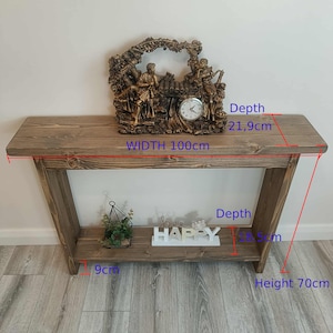 Op de afbeelding: Een rustieke houten console, 100 cm breed en 70 cm hoog, met een onderste plank. Het heeft een decoratieve klok met figuren en het woord "HAPPY". De diepte is 21,9 cm. De plankdiepte is 8,5 cm.