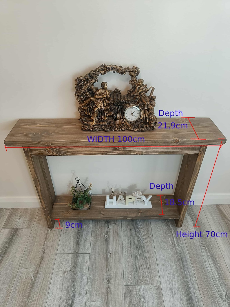Op de afbeelding: Een rustieke houten console, 100 cm breed en 70 cm hoog, met een onderste plank. Het heeft een decoratieve klok met figuren en het woord "HAPPY". De diepte is 21,9 cm. De plankdiepte is 8,5 cm.