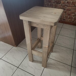 K&ouml;nnte beinhalten: Ein hellfarbener, quadratischer Holzstuhl mit rustikalem Design. Der Hocker hat vier stabile Beine und eine flache Sitzfl&auml;che. Die Holzmaserung ist sichtbar und verleiht ihm ein nat&uuml;rliches, unfertiges Aussehen. Geeignet als K&uuml;chen- oder Barhocker.