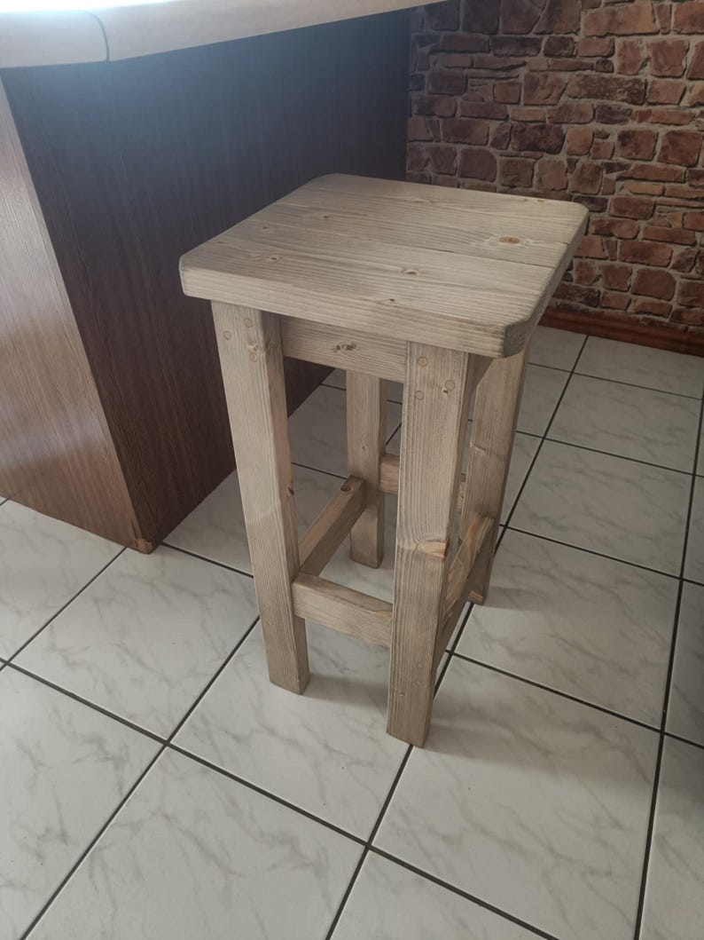 K&ouml;nnte beinhalten: Ein hellfarbener, quadratischer Holzstuhl mit rustikalem Design. Der Hocker hat vier stabile Beine und eine flache Sitzfl&auml;che. Die Holzmaserung ist sichtbar und verleiht ihm ein nat&uuml;rliches, unfertiges Aussehen. Geeignet als K&uuml;chen- oder Barhocker.