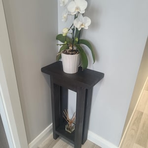 Op de afbeelding: Een donkergrijze houten plantenstandaard met een witte orchidee in een witte pot. De standaard heeft een rechthoekige bovenkant en een onderste plank. De orchidee heeft witte bloemen en groene bladeren. Een klein glazen potje met riet staat op de onderste plank.