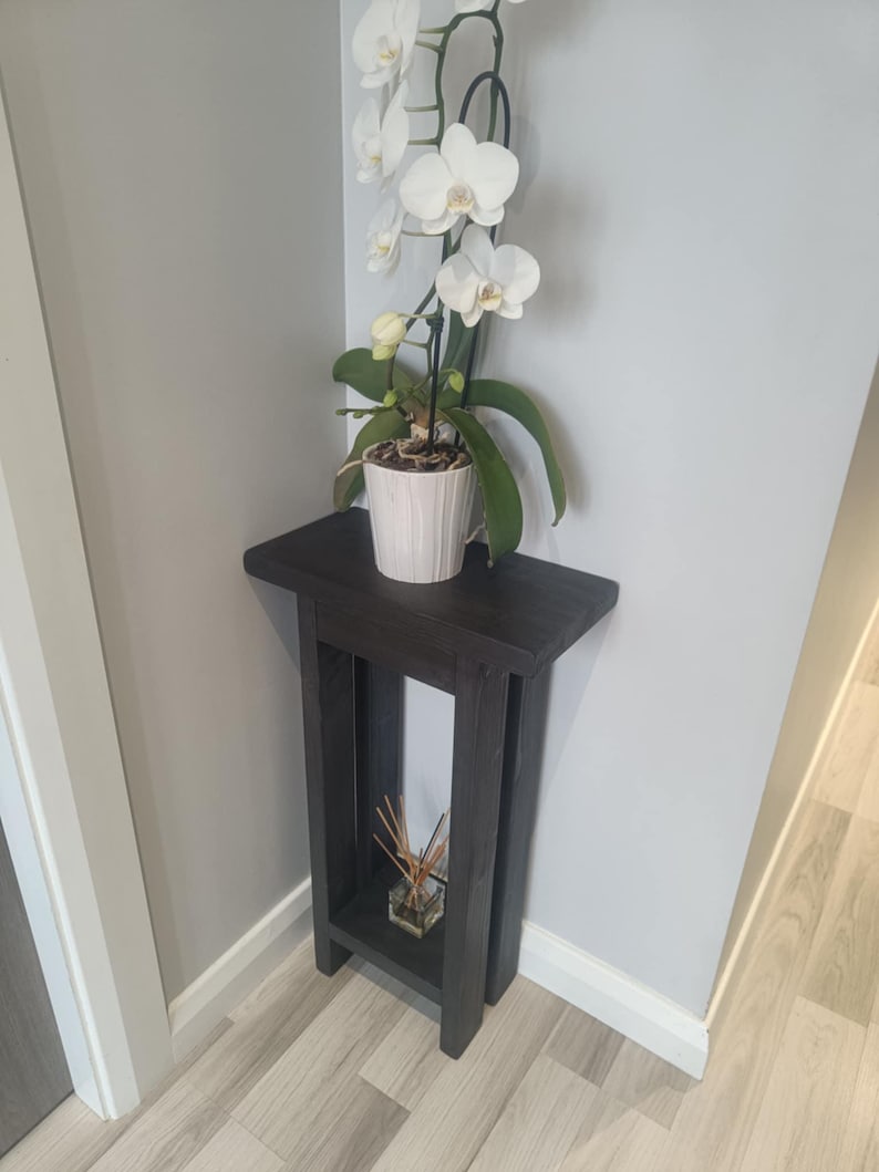 Op de afbeelding: Een donkergrijze houten plantenstandaard met een witte orchidee in een witte pot. De standaard heeft een rechthoekige bovenkant en een onderste plank. De orchidee heeft witte bloemen en groene bladeren. Een klein glazen potje met riet staat op de onderste plank.