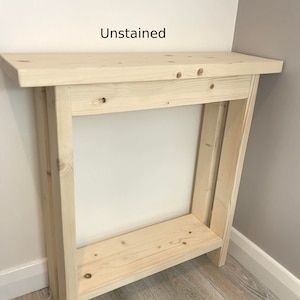 Op de afbeelding: Ongeschilderde houten console tafel. De tafel is gemaakt van lichtgekleurd hout en heeft een rechthoekig bovenblad, een onderste plank en vier poten. De tekst "Unstained" is zichtbaar op de muur achter de tafel.