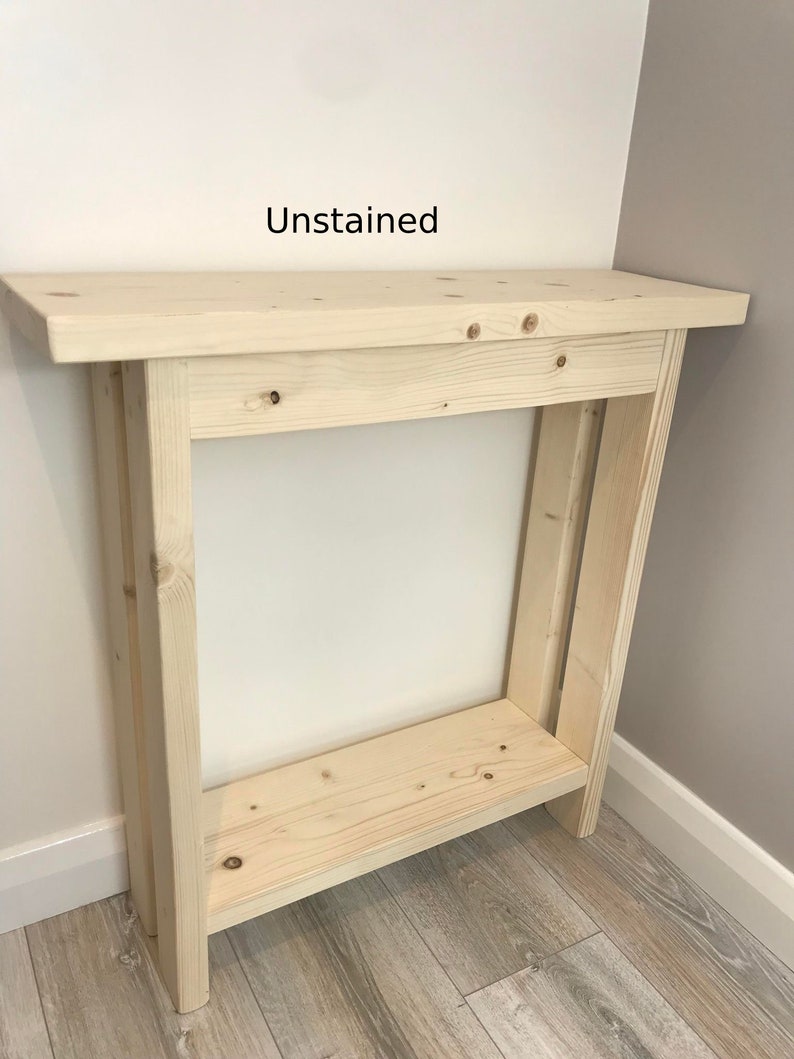 Op de afbeelding: Ongeschilderde houten console tafel. De tafel is gemaakt van lichtgekleurd hout en heeft een rechthoekig bovenblad, een onderste plank en vier poten. De tekst "Unstained" is zichtbaar op de muur achter de tafel.