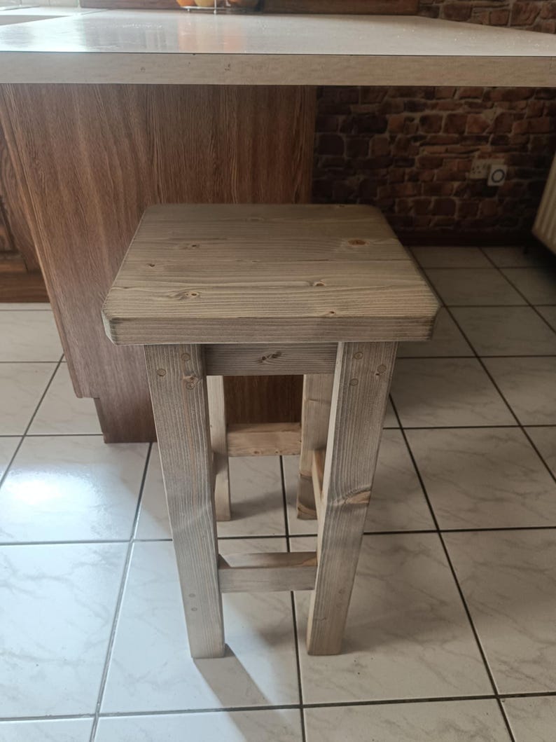 K&ouml;nnte beinhalten: Ein rustikaler, hell get&ouml;nter Holzstuhl mit quadratischer Sitzfl&auml;che und vier stabilen Beinen. Der Hocker ist mit sichtbarer Holzmaserung und Verbindungen versehen und zeigt eine nat&uuml;rliche, unbehandelte &Auml;sthetik. Ideal f&uuml;r K&uuml;chen oder Barbereiche.