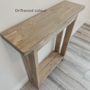 Op de afbeelding: Een houten console in een driftwood kleur. De tafel heeft een rechthoekig blad en een onderste plank, ondersteund door schuine poten. De tekst "Driftwood colour" is zichtbaar op de bovenkant van de tafel.