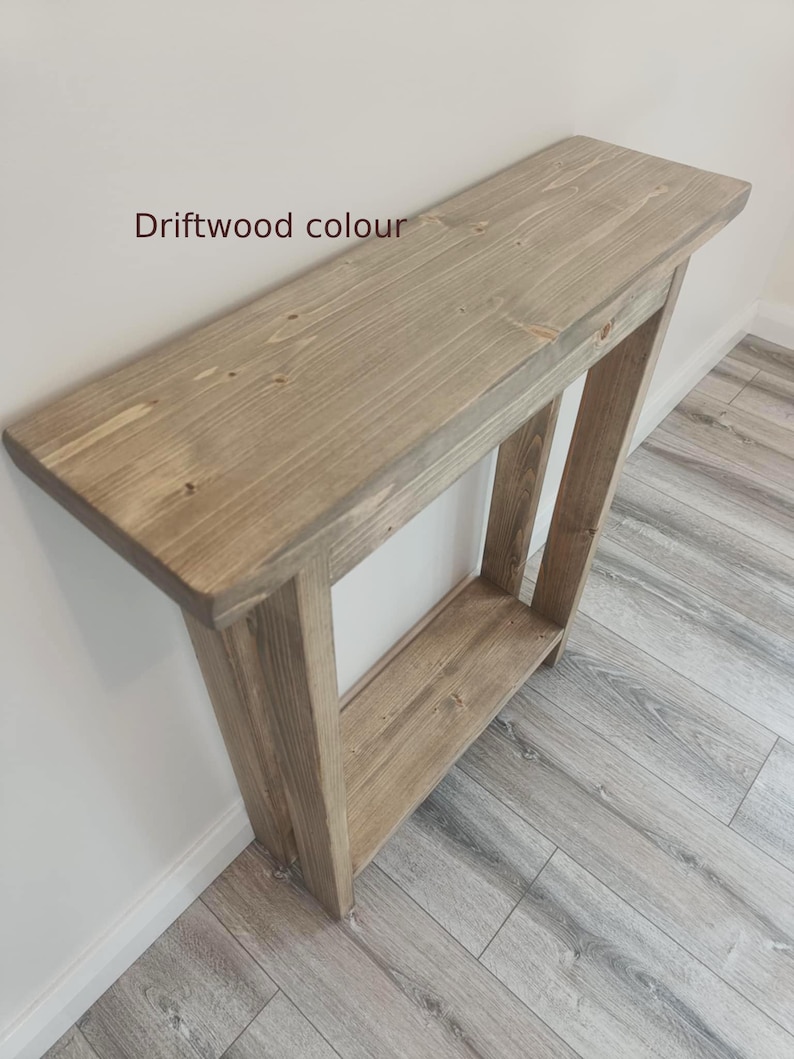 Op de afbeelding: Een houten console in een driftwood kleur. De tafel heeft een rechthoekig blad en een onderste plank, ondersteund door schuine poten. De tekst "Driftwood colour" is zichtbaar op de bovenkant van de tafel.