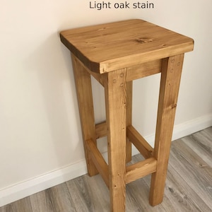 K&ouml;nnte beinhalten: Ein hell eichenfarbener Holzstuhl mit quadratischer Sitzfl&auml;che und vier stabilen Beinen. Der Text "Light oak stain" ist auf dem Bild zu sehen. Der Hocker ist aus Holz und hat eine nat&uuml;rliche Oberfl&auml;che.