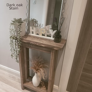 Op de afbeelding: Een donker eikenhouten console met een plank, met een witte vaas met gedroogde bloemen. Boven de tafel hangt een spiegel met een golvend zilveren frame. Het woord "HAPPY" staat op de tafel, samen met een plant in een pot en een diffuser.