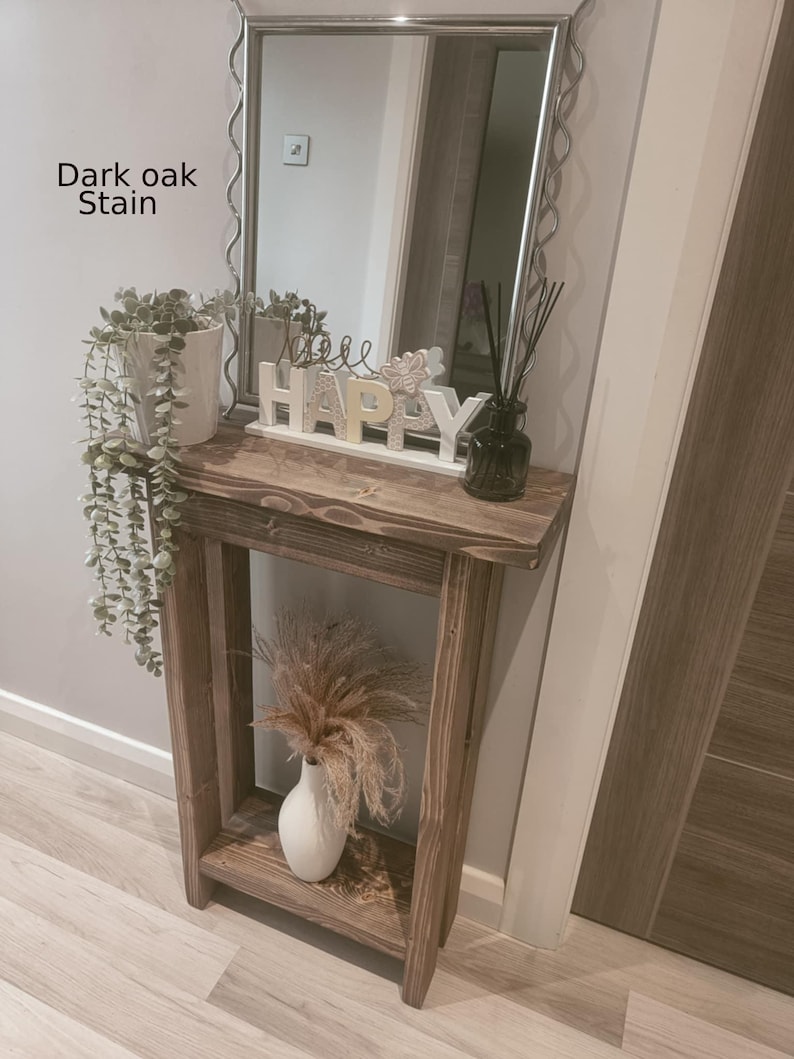 Op de afbeelding: Een donker eikenhouten console met een plank, met een witte vaas met gedroogde bloemen. Boven de tafel hangt een spiegel met een golvend zilveren frame. Het woord "HAPPY" staat op de tafel, samen met een plant in een pot en een diffuser.