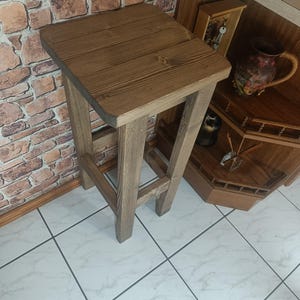 K&ouml;nnte beinhalten: Rustikaler Barhocker aus Holz mit quadratischer Sitzfl&auml;che und vier stabilen Beinen. Der Hocker ist aus hellbraunem Holz gefertigt, das die nat&uuml;rliche Maserung zeigt. Das Design ist schlicht und funktional, geeignet f&uuml;r K&uuml;che oder Bar.